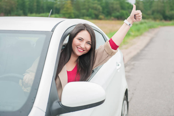 Belle jeune femme heureuse montrant pouces levés et assis dans la voiture blanche sur la route en plein air - Photo, image