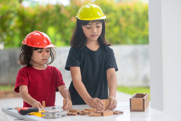 Aziatische broers en zussen meisjes dragen technische hoeden gebouw huis van het houten speelgoed. Om te leren en ontwikkeling te verbeteren, weinig architect. - Foto, afbeelding
