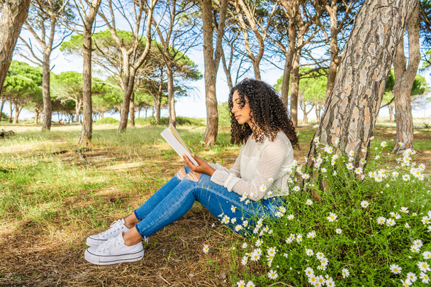 Екзотична чорна іспаномовна дівчинка сидить у сосновому лісі серед квітучих маргаритки, які читають улюблену романтичну книгу. Цікава молода афро-американська жінка проводить час в природі зі своєю пристрастю до любовних романів - Фото, зображення
