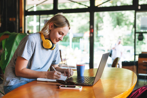 Young smiling blonde woman freelancer with yellow headphones working on notebook on the table at cafe. Girl student studying online course - Photo, image