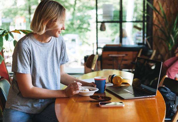 Giovane donna bionda sorridente freelance con cuffie gialle che lavora su notebook sul tavolo al caffè. Ragazza studentessa che studia sul corso online. Donna che fa colazione nel caffè - Foto, immagini