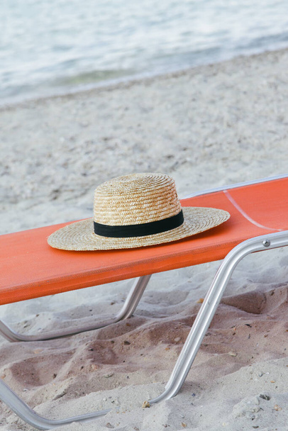 Ψάθινο καπέλο και πορτοκαλί ξαπλώστρα στην παραλία, χωρίς ανθρώπους. Ειδυλλιακή και γαλήνια έννοια καλοκαιρινές διακοπές. - Φωτογραφία, εικόνα
