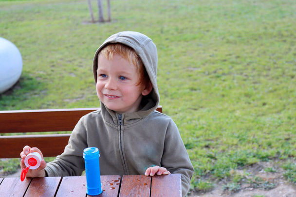 Un chico guapo de cuatro años con capucha mira a la distancia y sonríe. Niño feliz con burbujas de jabón.  - Foto, imagen