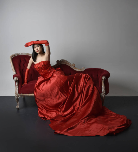 Portret pięknej młodej Azjatki w czerwonym gorsecie, długich rękawiczkach operowych i ozdobnym nakryciu głowy korony. Postawa siedząca z gestykulacyjnymi dłońmi odizolowanymi na tle studia światła. - Zdjęcie, obraz