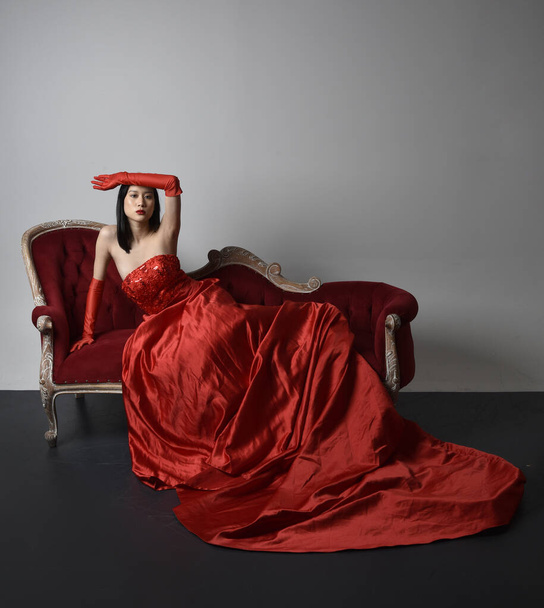 Full length  portrait of beautiful young asian woman wearing red corset, long opera gloves and ornate crown headdress. Sitting pose with gestural hands  isolated on light studio background. - Fotó, kép