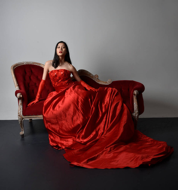 Teljes hosszúságú portré gyönyörű fiatal ázsiai nőről piros fűzővel, hosszú operakesztyűvel és díszes fejdísszel. Ülő póz geszturális kézzel elszigetelt fény stúdió háttér. - Fotó, kép