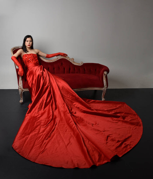 Portrait complet de belle jeune femme asiatique portant un corset rouge, de longs gants d'opéra et une coiffe de couronne ornée. Pose assise avec les mains gestuelles isolées sur fond de studio léger. - Photo, image