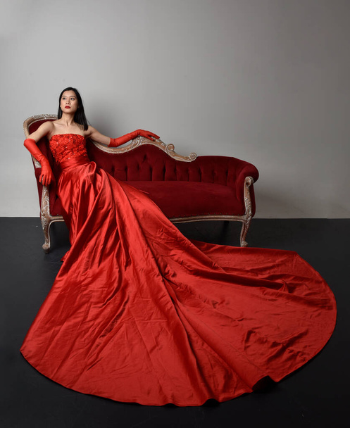 Täyspitkä muotokuva kaunis nuori aasialainen nainen yllään punainen korsetti, pitkä ooppera käsineet ja koristeellinen kruunu päähine. Istuminen aiheuttaa eleellinen kädet eristetty valo studio tausta. - Valokuva, kuva