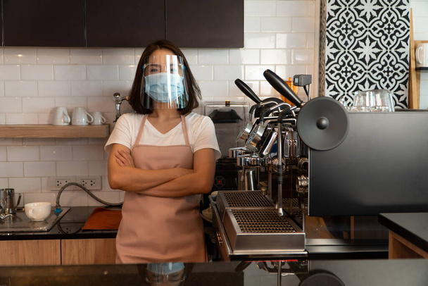 Yüz maskesi takan mutlu bir Asyalı garsonun portresi. Bir kafede duruyor. Küçük bir işletme sahibi ve bir kafe konseptiyle işe başlıyor. - Fotoğraf, Görsel