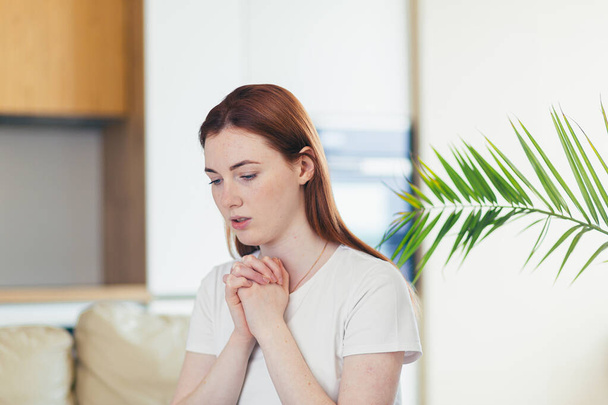 Closeup portrait of a young woman praying sincerely with folded arms at home or indoors. Sincere prayer of a Caucasian girl or female in the church with closed eyes alone. close up at home in the room - Foto, imagen