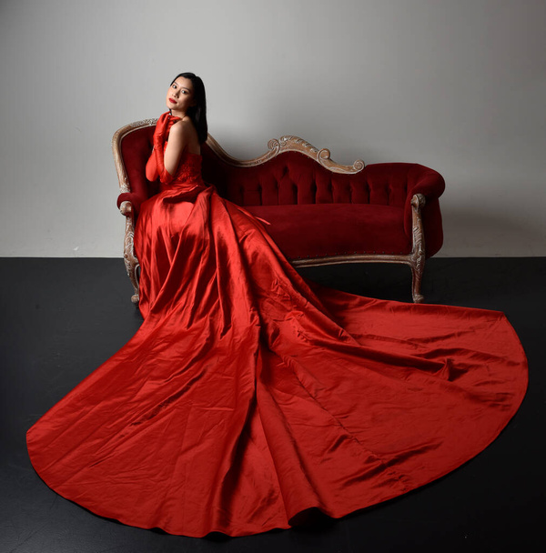 Portrait complet de belle jeune femme asiatique portant un corset rouge, de longs gants d'opéra et une coiffe de couronne ornée. Pose assise avec les mains gestuelles isolées sur fond de studio léger. - Photo, image