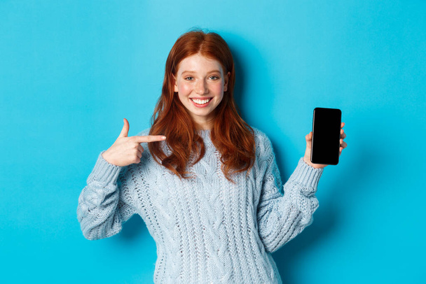 Zufriedenes rothaariges Mädchen zeigt auf den Handybildschirm, zeigt Smartphone-App oder Online-Promo und lächelt, steht im Pullover vor blauem Hintergrund - Foto, Bild