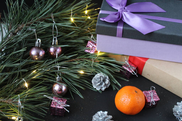New Year's Eve christmas background. Gifts boxes in a package with bows ribbons lie next to the spruce pine burning festive garland next to the mandarin cones silver on a black background. Christmas Eve festive atmosphere Christmas Eve. - Photo, Image