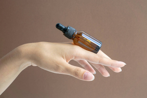 Mock-up of glass dropper bottle lies on woman's hand. Close-up, brown background, copy space. Concept of beauty industry. - Fotoğraf, Görsel