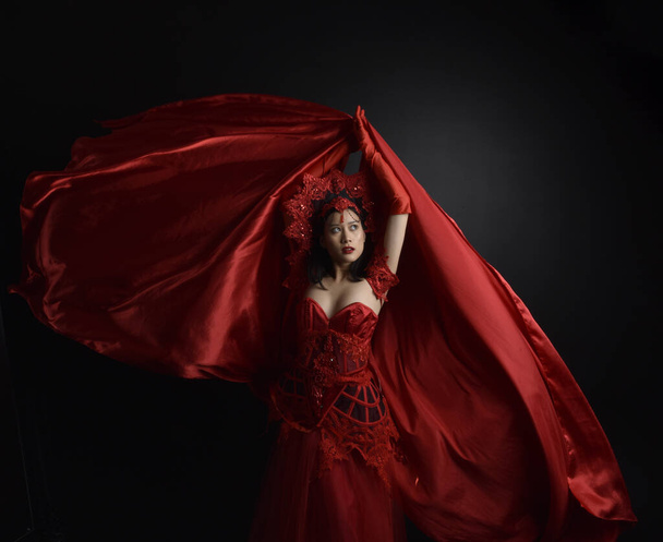 portret van een mooie jonge Aziatische vrouw met rood korset, lange operahandschoenen en sierlijke kroonhoofdtooi. Zittende houding met gebaren handen geïsoleerd op licht studio achtergrond. - Foto, afbeelding