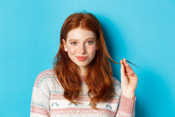 Gros plan d'une adolescente rousse confiante et grincheuse regardant la caméra avec plaisir, jouant avec des mèches de cheveux et souriant, debout sur un fond bleu - Photo, image