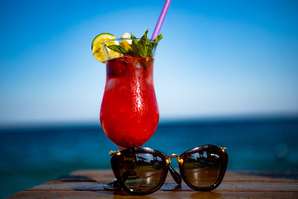 cóctel rojo con gafas de sol en la orilla del mar en una mesa de madera - Foto, Imagen