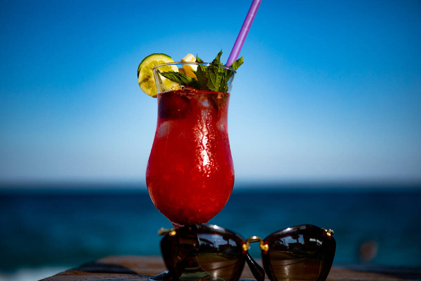 cóctel rojo con gafas de sol en la orilla del mar en una mesa de madera - Foto, Imagen