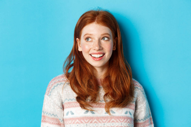 Close-up of dreamy redhead girl in winter sweater looking left, smiling and staring at promo offer, standing against blue background - Foto, immagini