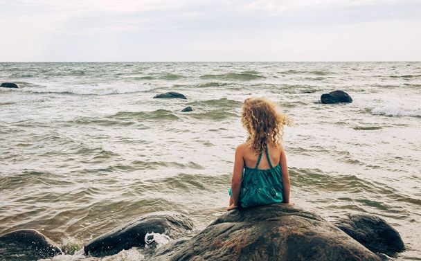 Enfant fille de 7 ans assis seul sur un gros rocher et regardant la mer agitée en été, ciel couvert. - Photo, image