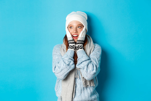 Возбужденная рыжая девушка смотрит налево на логотип, одетая в зимнюю одежду, шапочку, перчатки и свитер, стоя на синем фоне - Фото, изображение