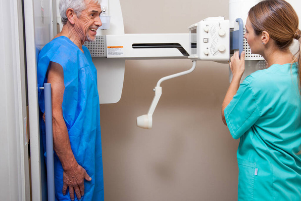 männliche Patientin in den 60ern, die sich einem Röntgentest unterzieht, unterstützt von einer lächelnden schönen Ärztin. Glückliches Krankenhauskonzept. - Foto, Bild