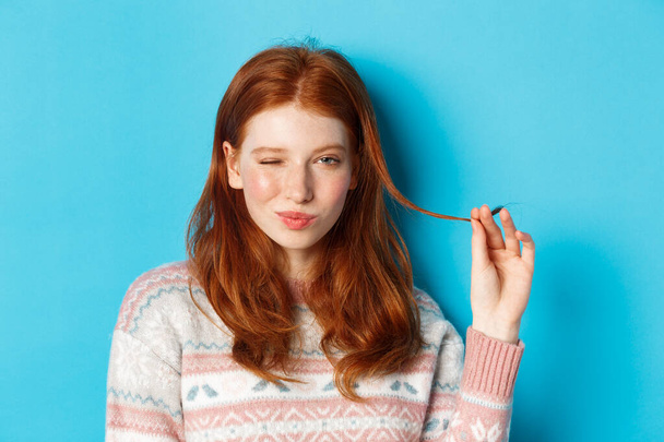 Gros plan de fille rousse effrontée jouant avec des mèches de cheveux, clignant des yeux et souriant à la caméra, debout sur fond bleu - Photo, image