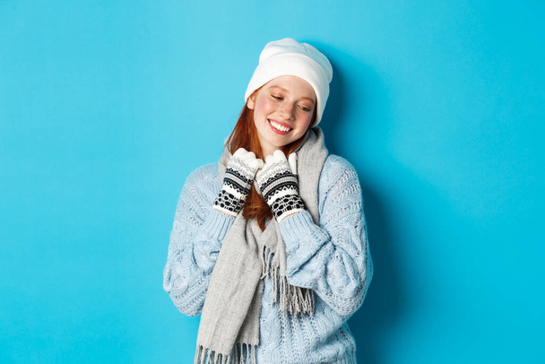 Zimní a prázdninový koncept. Roztomilé zrzka dívka červená a dívá se pryč zasněný, na sobě teplé beanie, svetr a šála s rukavicemi, stojící nad modrým pozadím - Fotografie, Obrázek
