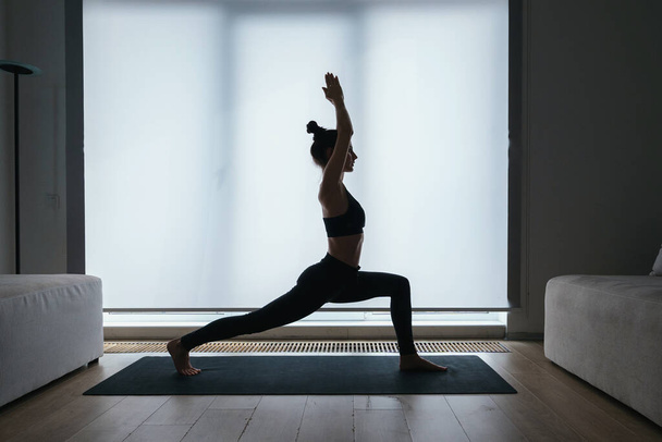 Silhouette of beautiful slim woman performing high crescent lunge yoga position while exercising at home, feeling calm and relaxed. Balance, zen and meditation concept - 写真・画像