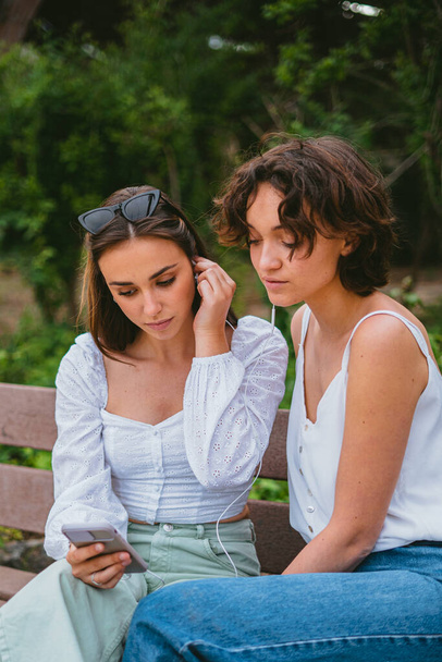 Dos adolescentes serias viendo un video mientras están sentadas en un banco de madera en el parque. - Foto, imagen