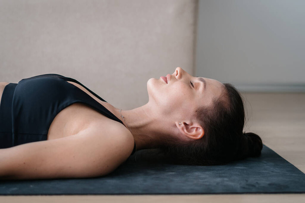 Attraktive Frau, die zu Hause meditiert, ruhig atmet und sich gleichzeitig auf ihre psychische Gesundheit, innere Harmonie und das Zen konzentriert. Junge Frau liegt beim Training auf Yogamatte - Foto, Bild