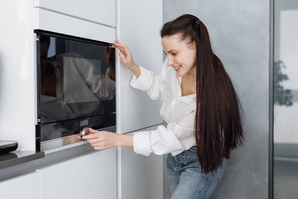 Gülümseyen genç bir kadının mikrodalga fırın topuzunu çevirmesi ve evdeki modern mobilyalı mutfağı ısıtırken zaman ve sıcaklık ayarlarını ayarlaması. - Fotoğraf, Görsel