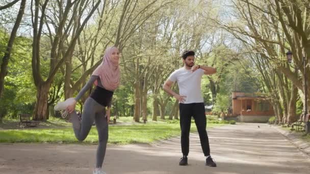 Активна жінка в спортивному одязі і хіджабі розтягує ноги в літньому парку. Молодий арабський чоловік стоїть на фоні і використовує розумний годинник. Спортивна активність на відкритому повітрі. Жінка розігрівається, поки чоловік використовує годинник
 - Кадри, відео