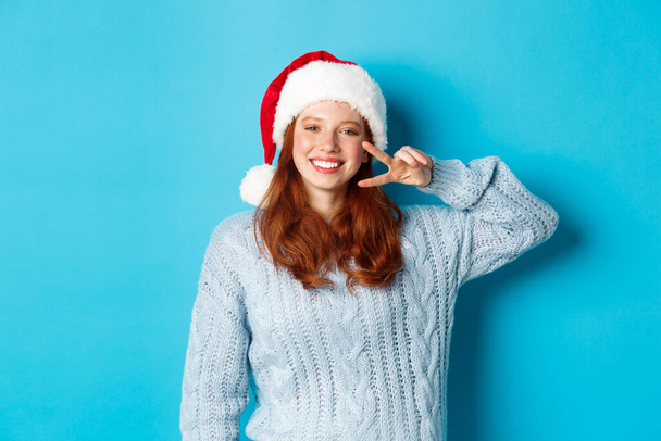 Winter holidays and Christmas Eve concept. Happy teenage girl with red hair, wearing santa hat, enjoying New Year, showing peace sign and smiling, standing over blue background - Photo, Image