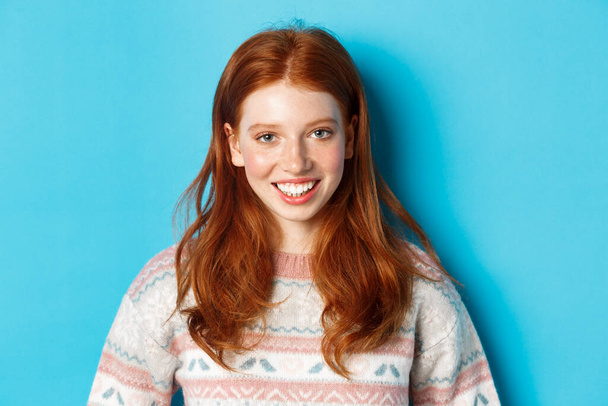 Close-up of cute redhead girl in sweater smiling happy at camera, standing against blue background - Foto, Imagem