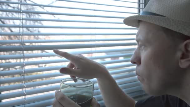 Een jonge man met een hoed met interesse onderzoekt wat er op straat gebeurt, drinkt uit een glas - Video