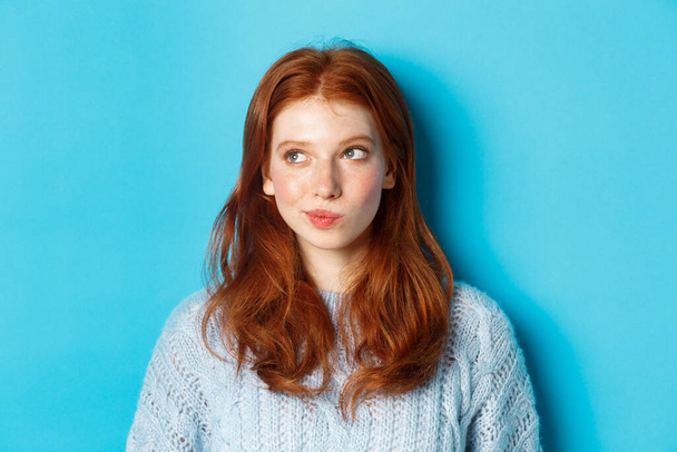 Close-up of cute redhead girl thinking and looking left, have an idea, standing against blue background - Foto, immagini