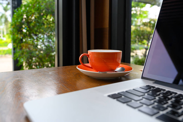 Copa de café capuchino caliente con portátil moderno en la mesa de madera dentro de la cafetería, enfoque selectivo en la taza de café. Espacio de trabajo empresarial, concepto de estilo de vida moderno - Foto, imagen