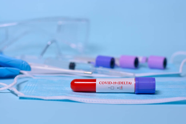 A test blood sample tube positive with delta variant or strain COVID-19. A test tube with a test result on a medical mask against the background of test tubes, thermometer, syringe, medical glasses - Foto, immagini