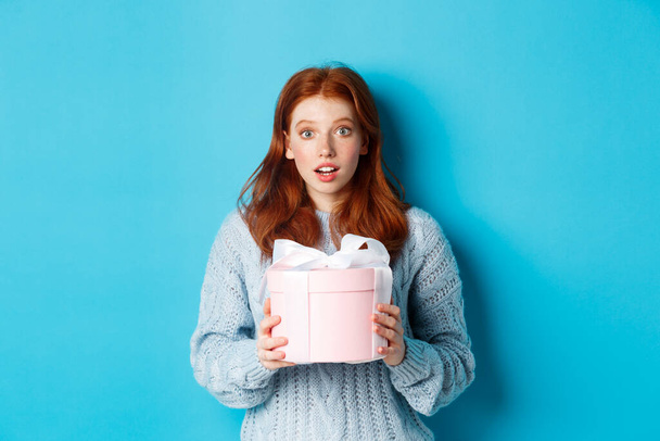 Surprised redhead girl receiving valentines gift, holding box with present and staring at camera amazed, wearing sweater, standing over blue background - Photo, Image