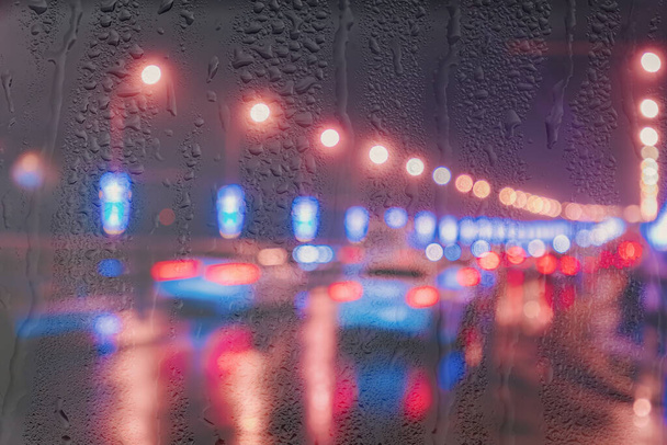 Blurry lights of moving cars and lanterns reflecting on the wet asphalt in the night city behind rain-covered window. Rainy bad autumn weather concept. - Foto, afbeelding