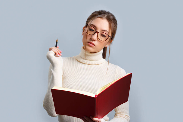 Pensive young woman looks into a notebook. Girl in glasses and with a pen on an isolated gray background. - Foto, Bild