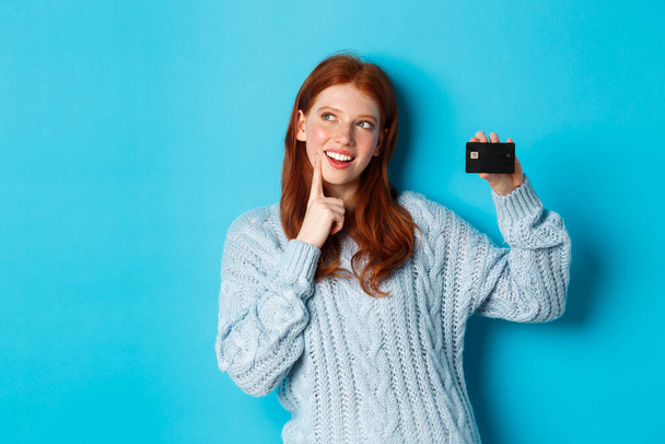 Image of thoughtful redhead girl thinking about shopping, showing credit card and pondering, standing over blue background - Photo, Image