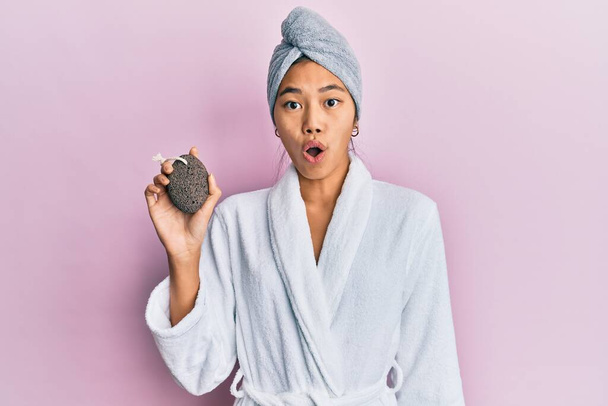 Młoda Chinka nosi czepek na ręczniki i szlafrok trzymając pumeks kamień przestraszony i zdumiony z otwartymi ustami dla zaskoczenia, niedowierzanie twarz  - Zdjęcie, obraz