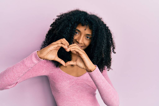 アフロの髪をしたアフリカ系アメリカ人女性は、ピンクのカジュアルなシャツを着て手でハートシンボルの形をするのが大好きです。ロマンチックなコンセプト.  - 写真・画像