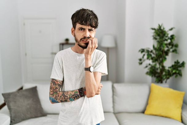 Spanyol férfi szakállal a nappaliban otthon stresszesnek és idegesnek tűnik, kezével a száján körmöket harap. szorongás.  - Fotó, kép