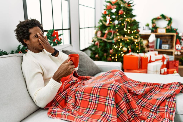 Joven afroamericano sentado en el sofá bebiendo café junto al árbol de navidad oliendo algo apestoso y asqueroso, olor intolerable, conteniendo la respiración con los dedos en la nariz. mal olor  - Foto, Imagen