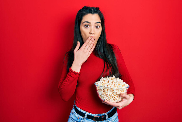 Young hispanic girl eating popcorn covering mouth with hand, shocked and afraid for mistake. surprised expression  - Foto, immagini