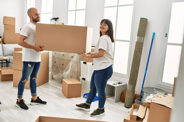 Молодая латиноамериканская пара улыбается счастливо держа большую картонную коробку в новом доме. - Фото, изображение