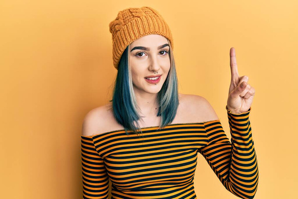 Mladá moderní dívka nosí vlněný klobouk ukazuje a ukazuje prstem číslo jedna a zároveň se usmívá sebevědomě a šťastně.  - Fotografie, Obrázek